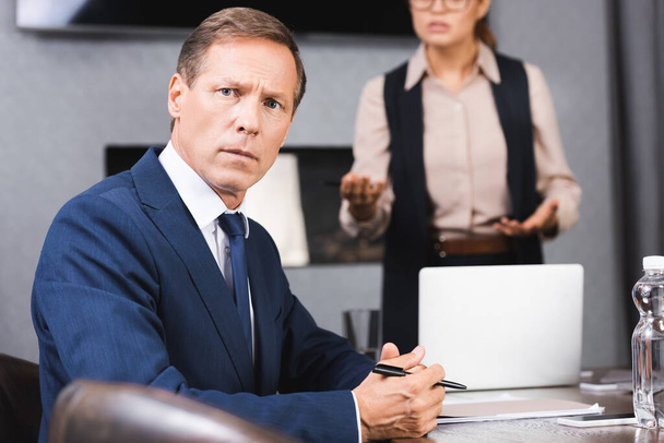 Scared businessman looking at camera while sitting at workplace with blurred female executive on background - Foto, imagen