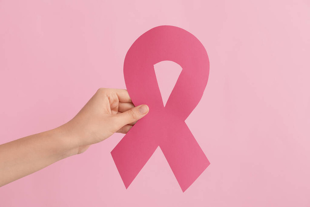 Γυναικείο χέρι με χάρτινη ροζ κορδέλα σε φόντο χρώματος. Έννοια ευαισθητοποίησης του καρκίνου του μαστού - Φωτογραφία, εικόνα