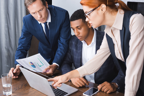 Бизнесмен, указывающая пальцем на ноутбук рядом с мультикультурными бизнесменами на рабочем месте - Фото, изображение