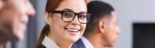 Happy redhead businesswoman in eyeglasses looking at camera with blurred african american man on background, banner - Photo, Image