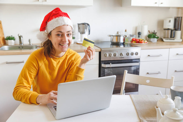 Nő a Mikulás sapkában online vásárol és aranykártyával fizet. Fiatal lány laptoppal karácsonyi ajándékot vásárol az interneten, a konyhában. Online karácsonyi vásárlási koncepció - Fotó, kép