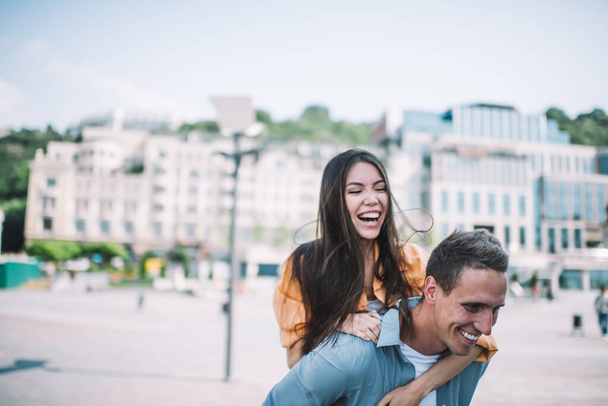 Handsome young man giving piggy back to joyful laughing girlfriend while walking together and enjoying romantic city tour on summer day - Valokuva, kuva