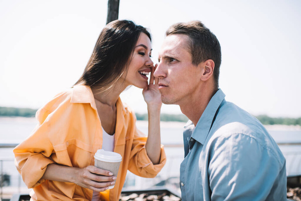 Laughing Asian young female whispering in ear of focused best friend sharing with secret while having coffee together sitting on promenade on sunny day - Foto, immagini