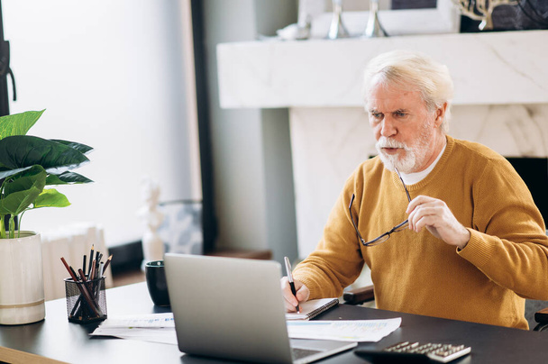Mature gray-haired man is looking surprised at laptop screen while sitting and using laptop at home office - Fotoğraf, Görsel