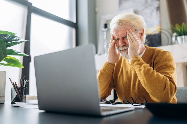 Älterer, müder, grauhaariger Mann, der vor Kopfschmerzen die Augen schließt und müde von der langen Arbeit im Laptop ist - Foto, Bild