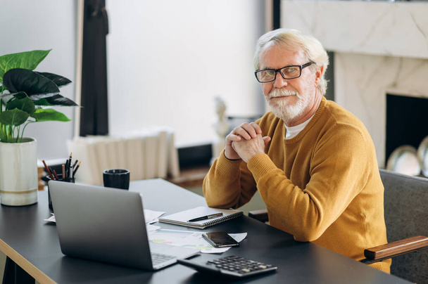 Літній сіроволосий чоловік в окулярах сидить за своїм робочим столом у своєму домашньому офісі і дивиться в камеру
 - Фото, зображення