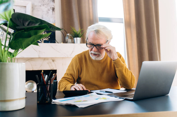Зрелый седой предприниматель в очках работает дистанционно дома с помощью калькулятора для ноутбука и документов, сидя дома за рабочим столом - Фото, изображение
