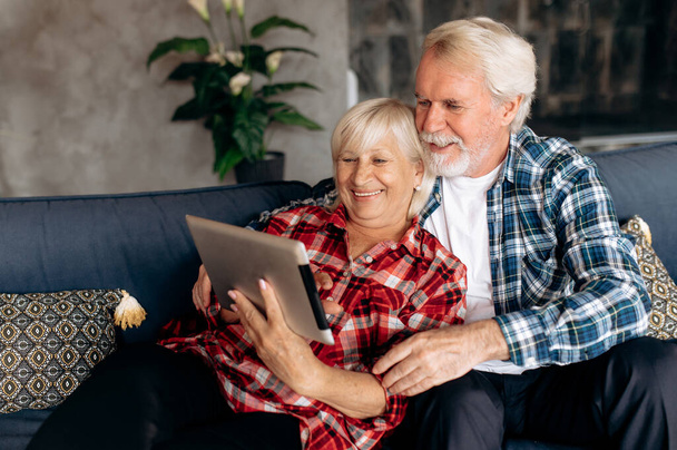 Pareja de ancianos modernos. Los cónyuges sonrientes maduros pasan su tiempo libre en casa en el sofá en una red social usando una tableta - Foto, Imagen