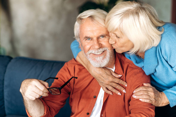 Ευτυχισμένο ηλικιωμένο γκριζομάλλικο ζευγάρι αγκαλιάζεται και κάθεται στον καναπέ στο σπίτι, η σύζυγος φιλάει τον άντρα και ο σύζυγος κοιτάζει την κάμερα και χαμογελάει - Φωτογραφία, εικόνα