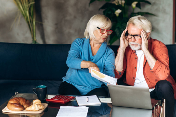 Online bill payment. Mature sad retirees count and pay their utility bills using calculator and laptop while sitting on a sofa at home - Photo, Image