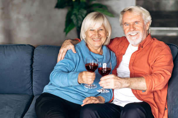 Happy starszych kaukaski para relaks w domu na kanapie picia wina przytulanie i patrząc w aparat z przyjemnym uśmiechem - Zdjęcie, obraz