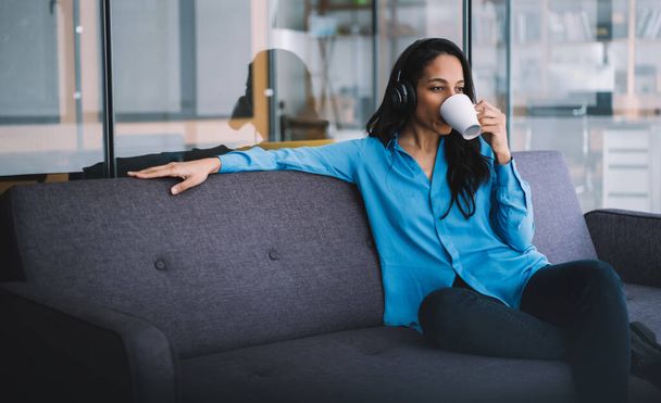 Relaxed ethnic female entrepreneur in headphones dressed in formal suit drinking coffee while sitting on couch at work and looking away - Fotografie, Obrázek
