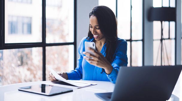 Attractive African American adult business lady in formal clothes holding smartphone and concentrating on documents while sitting at round white table with digital tablet and laptop near big window in office during daytime - Foto, Imagem