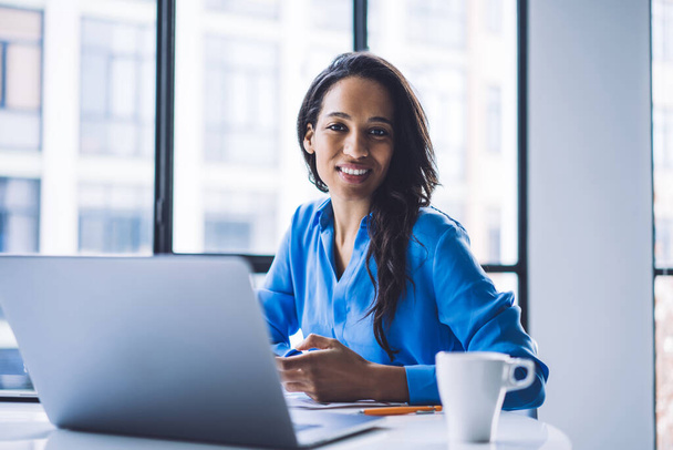 Mujer afroamericana sonriente positiva en ropa corporativa usando el ordenador portátil mientras está sentado en la mesa con la taza de café en la oficina moderna luz - Foto, imagen
