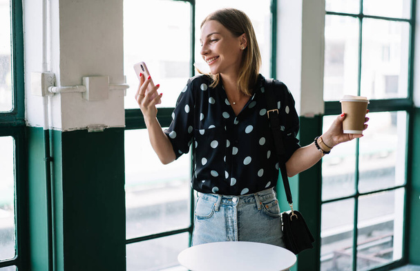 Positive millennial woman watching funny video on mobile phone enjoying coffee break in cafeteria, smiling carefree hipster girl millennial checking messages and mail via application on smartphone - Foto, Bild