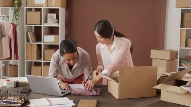 Tracking of multi-ethnic women checking order in office of small online store. Asian woman taking garments out of cardboard box as her black female colleague writing notes and looking at laptop - Footage, Video