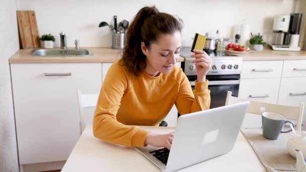 Een vrouw die online winkelt en betaalt met een gouden creditcard. Jong meisje zit met laptop kopen op internet voer credit card gegevens op keuken indoor achtergrond. Online winkelen e-commerce concept - Video