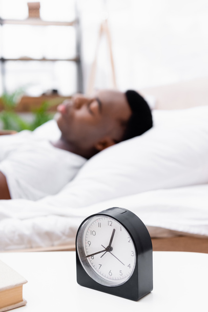 Clock and book on bedside table near african american man sleeping on blurred background  - Фото, зображення
