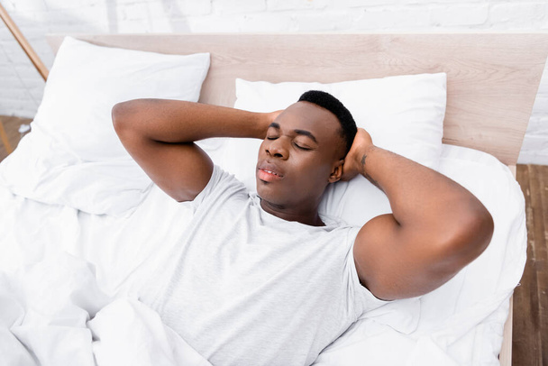 Hombre afroamericano con las manos cerca de la cabeza acostado en la cama por la mañana  - Foto, imagen