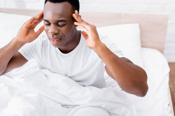 Homme afro-américain aux yeux fermés souffrant de maux de tête au lit sur fond flou  - Photo, image