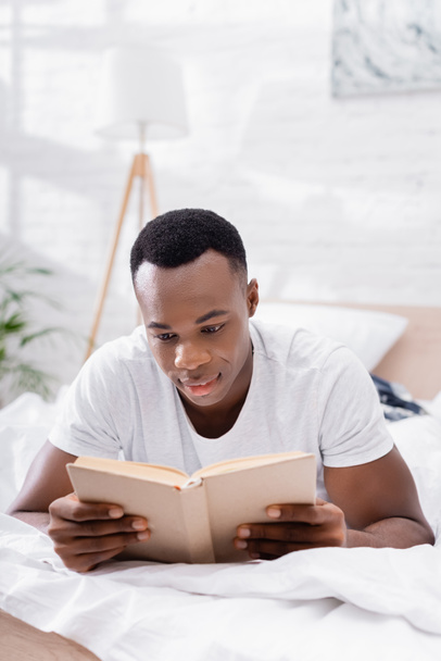 African american man reading book while lying on bed at morning  - 写真・画像