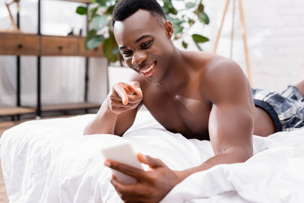 Alegre afroamericano hombre apuntando al teléfono inteligente en primer plano borroso en la cama  - Foto, Imagen