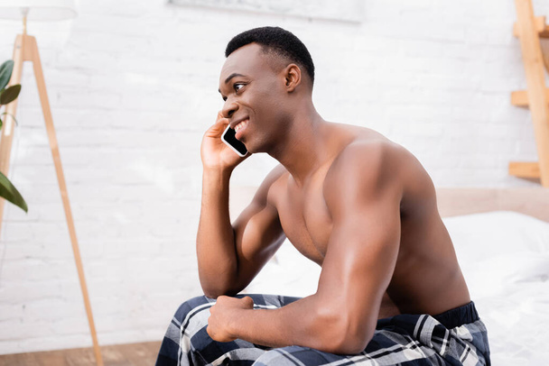 Mosolygós afro-amerikai férfi kockás nadrágban beszél okostelefonon az ágyban  - Fotó, kép