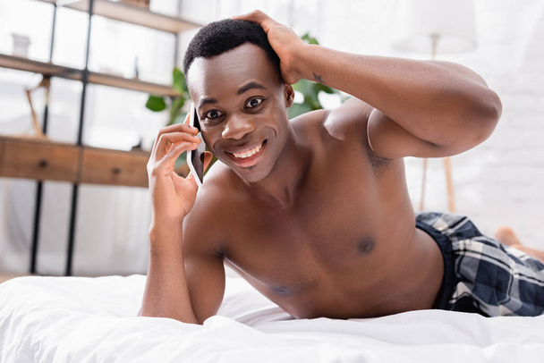 Mosolygós afro-amerikai férfi beszél okostelefonon az ágyon otthon  - Fotó, kép