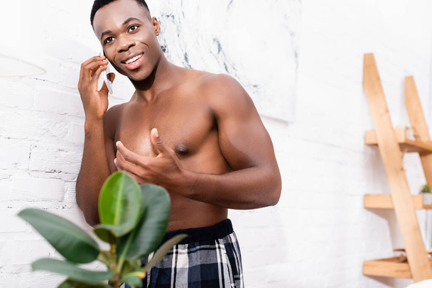 Félmeztelen afro-amerikai férfi beszél okostelefonon közel növény elmosódott előtérben otthon  - Fotó, kép