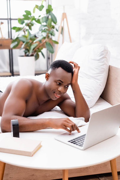 Shirtless african american man using laptop near clock and book on coffee table in bedroom  - 写真・画像