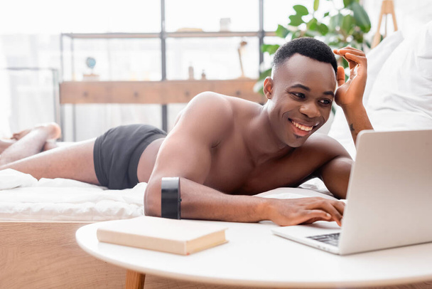 Африканський американець у штанах з ноутбуком під час лежання на ліжку біля книжки і годинника на розмитому передньому плані.  - Фото, зображення