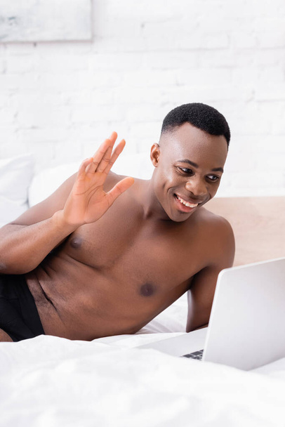Shirtless african american man having video call on laptop on bed at morning  - Photo, Image