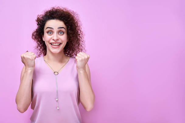 Portrait of armenian positive curly haired girl in pink dress, expressive face with bulging eyes and smile, clenched fists celebrating victory. Studio shot isolated on wall background - Photo, image