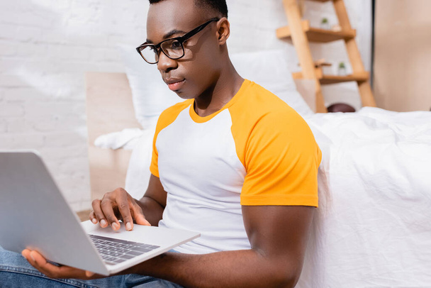 African american freelancer in eyeglasses using laptop on blurred foreground in bedroom  - 写真・画像