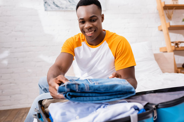 Smiling african american man putting jeans in suitcase on blurred foreground at home  - Photo, Image