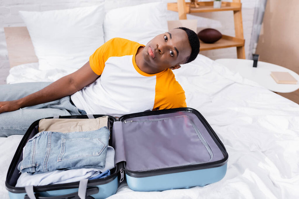 Suitcase with clothes near african american man looking at camera on bed  - Photo, Image