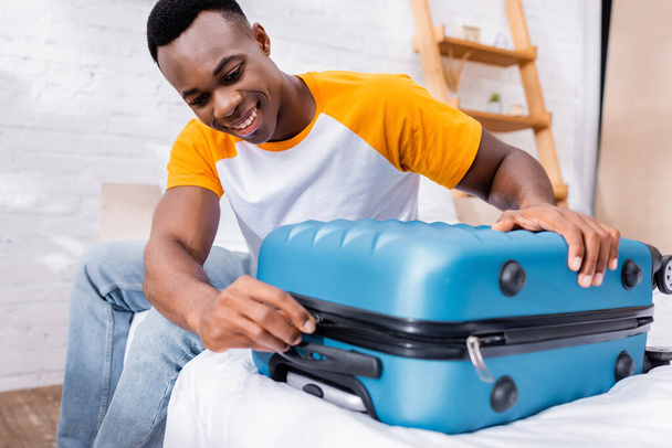 Positive african american man locking suitcase on blurred foreground on bed  - Фото, зображення