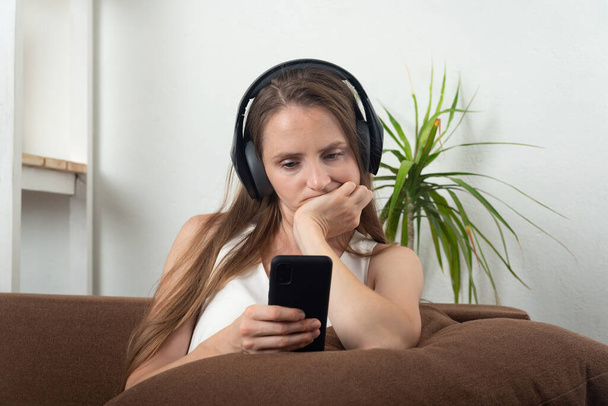 Νεαρή γυναίκα με ακουστικά κοιτάζει προσεκτικά στο smartphone της. Μαθήματα ακουστικής κατάρτισης. - Φωτογραφία, εικόνα