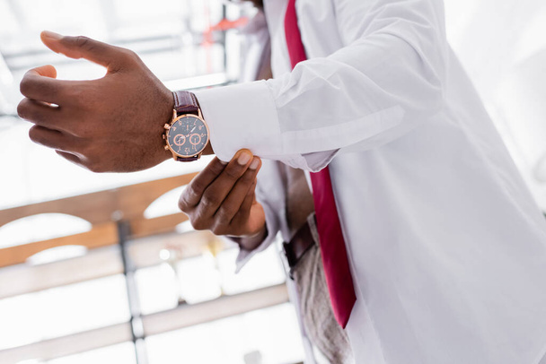 Vista recortada del reloj de pulsera en el brazo del hombre de negocios afroamericano con camisa blanca sobre fondo borroso  - Foto, Imagen