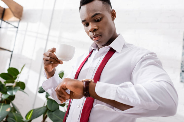 Αφροαμερικάνος επιχειρηματίας που κρατά την κούπα του καφέ και κοιτάζει το ρολόι χειρός σε θολή πρώτο πλάνο στο σπίτι  - Φωτογραφία, εικόνα