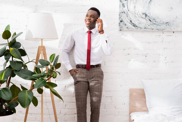 Glimlachende Afrikaanse Amerikaanse zakenman aan het praten op smartphone in de buurt van vloerlamp in de slaapkamer  - Foto, afbeelding