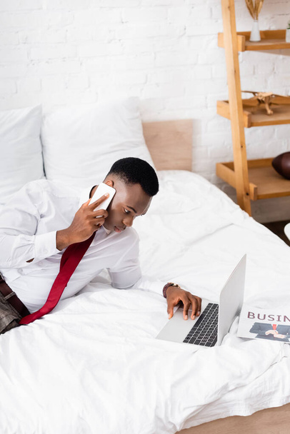 African american businessman using laptop while talking on smartphone near newspaper on bed  - Foto, afbeelding