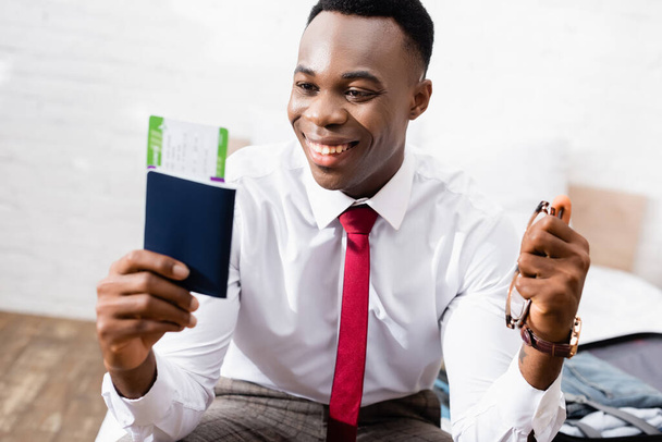 Homme d'affaires afro-américain souriant tenant lunettes et passeport avec billet d'avion au premier plan flou  - Photo, image