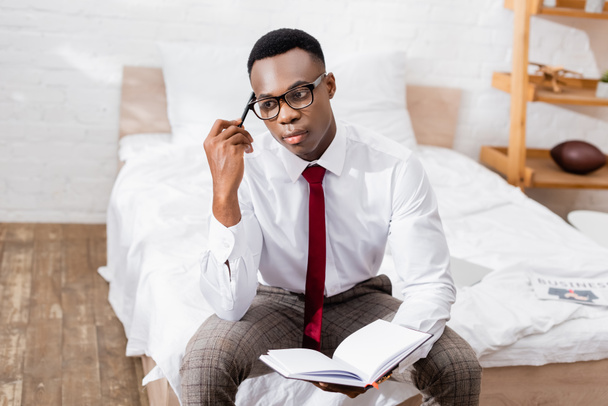 Pensive african american businessman in eyeglasses holding pen and notebook on bed on blurred background  - Fotografie, Obrázek