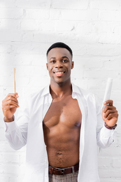 Muscular african american man in shirt smiling while holding toothpaste and toothbrush at home  - Foto, Imagen