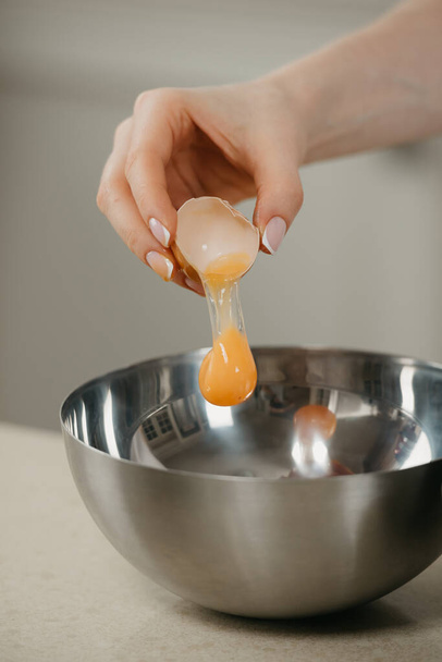 Una foto cercana de la mano de una niña que está dejando caer la yema del huevo orgánico de granja en el plato de sopa de acero inoxidable en una cocina. Una yema que cae. - Foto, imagen