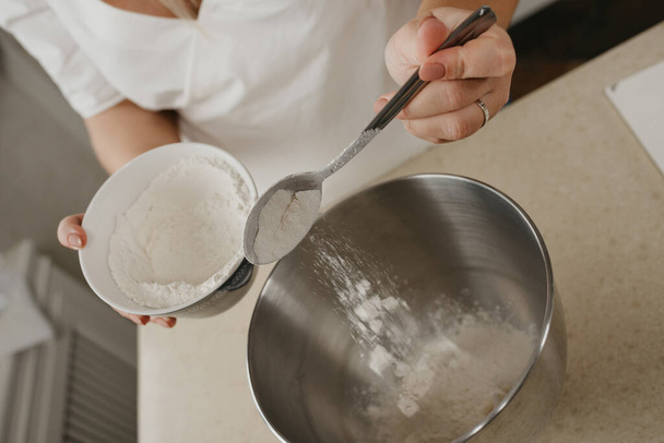 A close photo of the falling wheat flour from the spoon in the hand of the young woman into the stainless steel bowl of the mixer. - Photo, image