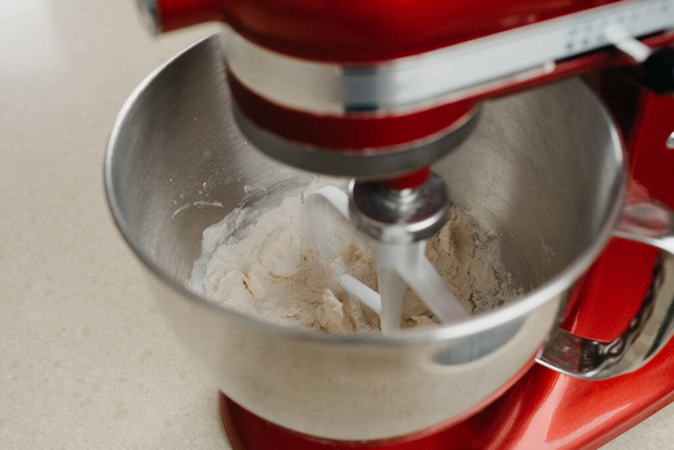 Een hechte foto van een rode standmixer die boter, amandelmeel en tarwemeel in een roestvrijstalen mengkom in een keuken klopt. - Foto, afbeelding
