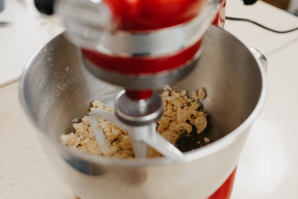 Een hechte foto van een rode standmixer die boter, amandelmeel, tarwebloem, poedersuiker en dooier in een roestvrijstalen mengkom in een keuken klopt. - Foto, afbeelding