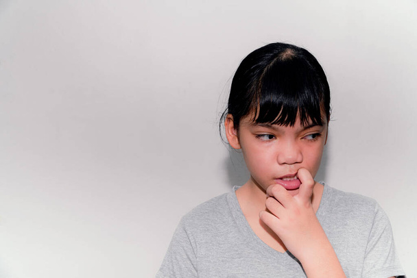 Egy ázsiai lány beleharap egy szögbe, és ideges valamiért. A szürke fal háttér, a probléma, hogy a mai gyerekek rágják a körmüket, talán személyes problémák vagy társadalmi félreértések miatt.. - Fotó, kép